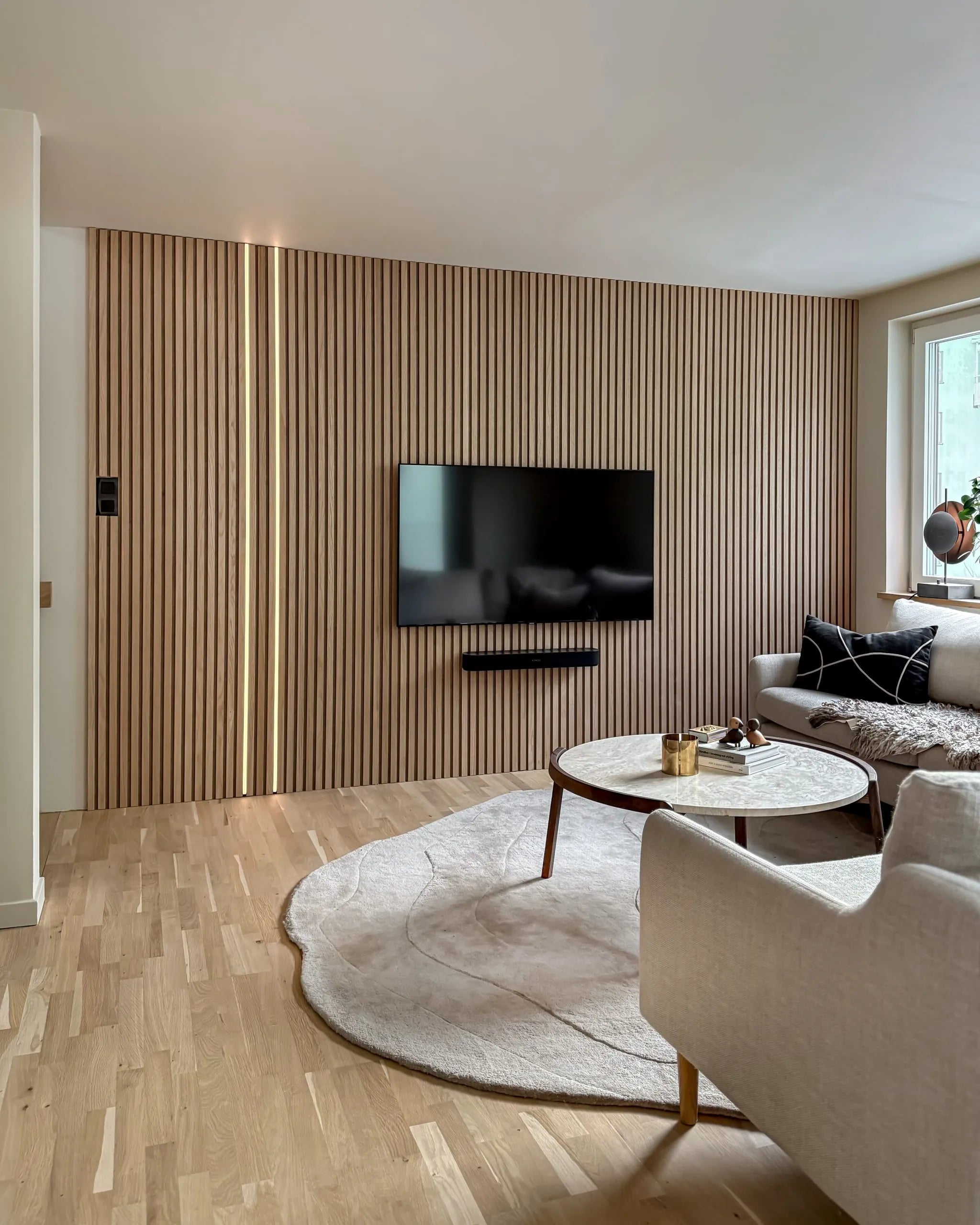 Modern living room with a Nordic Akupanel acoustic wood slat accent wall, mounted TV, round coffee table, cream sofa, large rug on light wood floors.